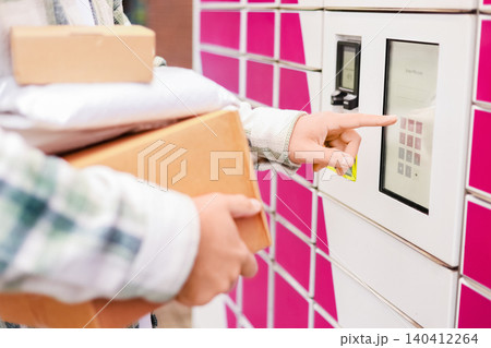 Person holding packages and entering a PIN on a touchscreen at a pink automated parcel locker. Convenient contactless delivery, e-commerce shopping, and logistics service. Person holding packages and entering a PIN on a touchscreen at a pink automated parcel locker. Convenient contactless delivery, e-commerce shopping, and logistics service. 140412264
