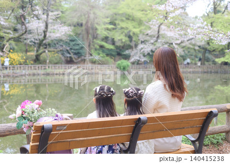 桜咲く公園のベンチで春を楽しむ母親と姉妹の後ろ姿 桜咲く公園のベンチで春を楽しむ母親と姉妹の後ろ姿 140415238