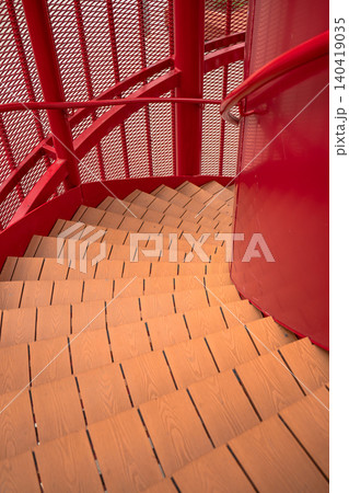 Looking down a vibrant red industrial spiral staircase featuring wooden steps and a mesh safety screen Looking down a vibrant red industrial spiral staircase featuring wooden steps and a mesh safety screen 140419035