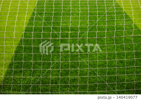 Close-up detail of a white soccer goal net grid against a blurred green grass field background Close-up detail of a white soccer goal net grid against a blurred green grass field background 140419077