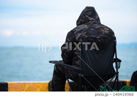 Rear view of a person wearing a camo hoodie sitting on a folding chair at a harbor pier 140419368