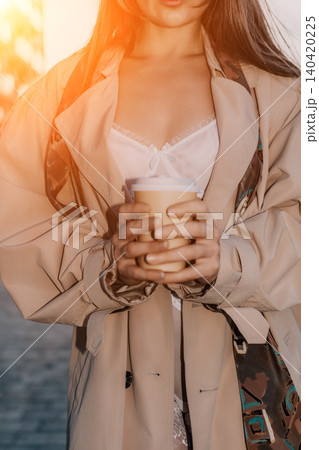 Coffee woman trench coat stylish person in a beige coat holds a hot drink in a paper cup while standing outdoors in golden light Coffee woman trench coat stylish person in a beige coat holds a hot drink in a paper cup while standing outdoors in golden light 140420225