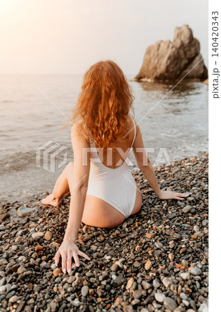 Woman Beach Sunset sitting on a pebble shore in a white swimsuit looking at the ocean waves during golden hour Woman Beach Sunset sitting on a pebble shore in a white swimsuit looking at the ocean waves during golden hour 140420343