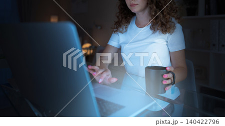 Woman working on laptop at night while holding a mug in soft light Woman working on laptop at night while holding a mug in soft light 140422796