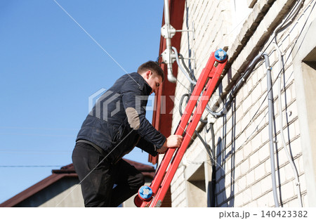 Worker climbing ladder on a house wall for installation or repair. Side view of young technician or electrician fixing cables outside building, house maintenance and telecommunication 140423382