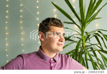 Man smiling and looking away indoors with garland lights on background. Portrait of happy young male sitting by a yucca palm plant, festive cozy atmosphere, lifestyle and human emotions concept Man smiling and looking away indoors with garland lights on background. Portrait of happy young male sitting by a yucca palm plant, festive cozy atmosphere, lifestyle and human emotions concept 140423383