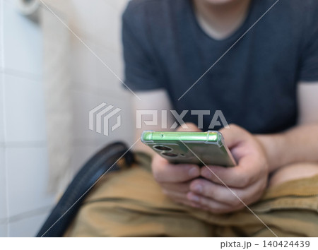 Man sitting on the toilet with a smartphone in hands. Guy scrolling phone on the loo, concept of prolonged sitting risk, screen addiction, and hemorrhoids. 140424439