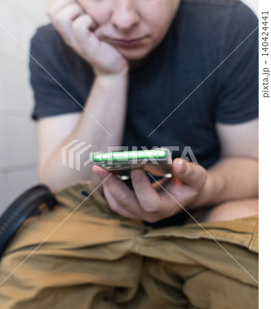Man sitting on the toilet with a smartphone in hands. Guy scrolling phone on the loo, concept of prolonged sitting risk, screen addiction, and hemorrhoids. Man sitting on the toilet with a smartphone in hands. Guy scrolling phone on the loo, concept of prolonged sitting risk, screen addiction, and hemorrhoids. 140424441