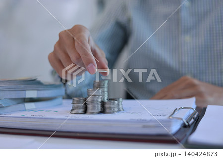Businessman carefully stacking silver coins on top of documents that feature various business charts and data visualizations. financial planning or investment tracking concept. Businessman carefully stacking silver coins on top of documents that feature various business charts and data visualizations. financial planning or investment tracking concept. 140424873