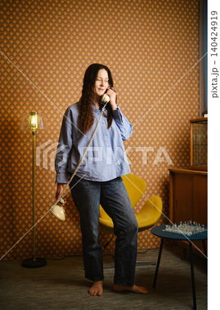 Woman speaking on rotary phone while standing in retro room with chess set Woman speaking on rotary phone while standing in retro room with chess set 140424919