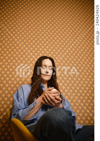 Woman sitting on chair holding cup in front of textured wall in indoor space Woman sitting on chair holding cup in front of textured wall in indoor space 140424926