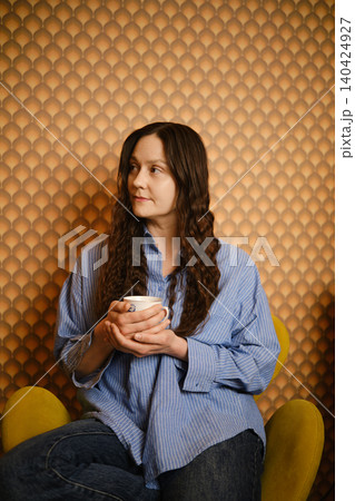 Woman in blue shirt sits in chair holding coffee cup in modern cafe with patterned wall Woman in blue shirt sits in chair holding coffee cup in modern cafe with patterned wall 140424927