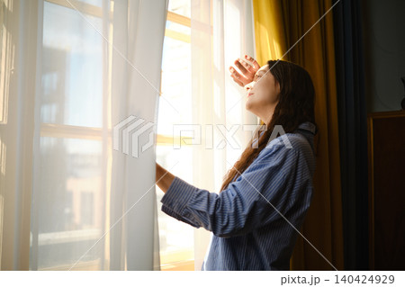 Woman looking out window and enjoying sunlight in a cozy indoor space during morning hours 140424929