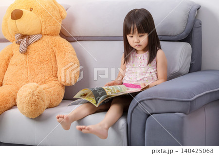 A giant teddy bear accompanies an Asian little girl as she reads on the sofa, a heartwarming scene of family learning and childhood ideals. 140425068