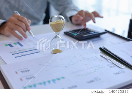 Close up of businessman analyzing growth charts and marketing statistics in office with hourglass on desk. Time management and financial analysis concept. 140425835