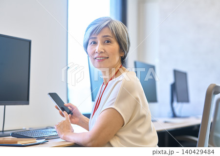 スマホを持ってデスクで振り向くグレーヘア女性 スマホを持ってデスクで振り向くグレーヘア女性 140429484