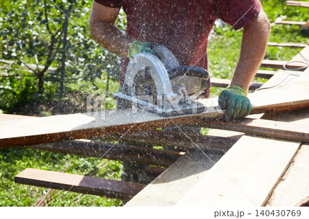 Worker using a circular saw to cut wooden boards outdoors, with sawdust flying as part of construction Worker using a circular saw to cut wooden boards outdoors, with sawdust flying as part of construction 140430769