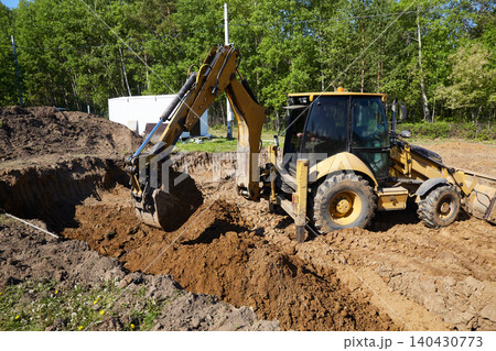 Backhoe loader digging and moving soil in a trench at a construction site, shaping the ground for foundation Backhoe loader digging and moving soil in a trench at a construction site, shaping the ground for foundation 140430773