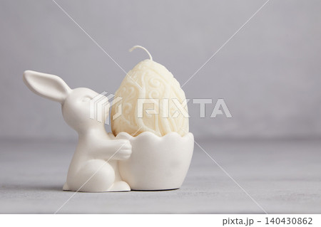A white Easter egg-shaped candle in a bunny-shaped holder against a gray background A white Easter egg-shaped candle in a bunny-shaped holder against a gray background 140430862
