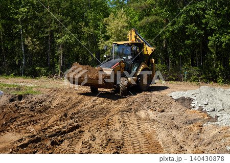 Backhoe loader carrying and dumping soil at a construction site, shaping ground and preparing foundation area 140430878