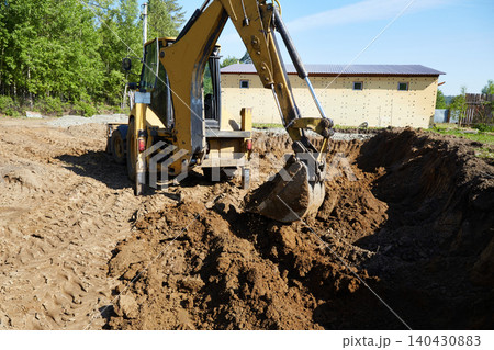 Backhoe excavator digging and moving soil in a trench at a residential construction site, preparing ground for foundation work. 140430883