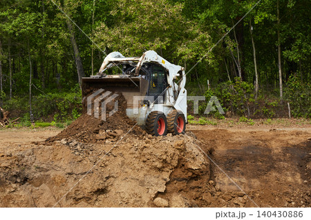 Compact skid steer loader pushing and dumping soil on a construction site near a forest Compact skid steer loader pushing and dumping soil on a construction site near a forest 140430886