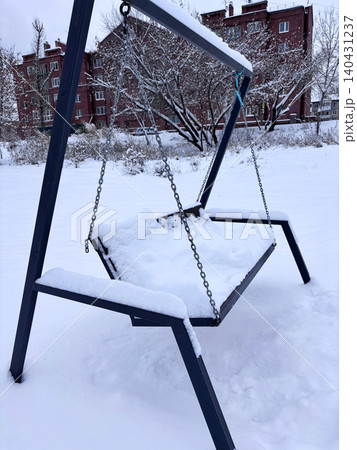 A swing sits in a park, blanketed with snow. Nearby buildings are visible, and trees are also covered in white. A swing sits in a park, blanketed with snow. Nearby buildings are visible, and trees are also covered in white. 140431237
