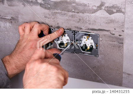 An electrician uses a screwdriver to install an electrical outlet on a wall covered with porcelain stoneware An electrician uses a screwdriver to install an electrical outlet on a wall covered with porcelain stoneware 140431255