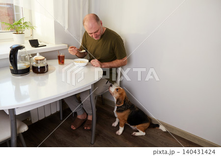 A man eats breakfast at the kitchen table, eating from a bowl while his dog sits nearby. A man eats breakfast at the kitchen table, eating from a bowl while his dog sits nearby. 140431424