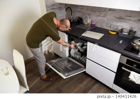 A man opens the dishwasher to unload clean dishes. A kitchen interior with white cabinets and wood floors A man opens the dishwasher to unload clean dishes. A kitchen interior with white cabinets and wood floors 140431426