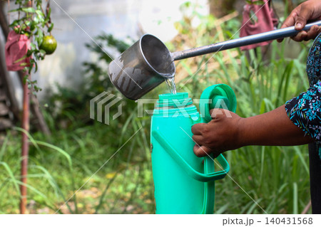 Weathered hands pouring steaming hot water from metal ladle into mint green thermos flask in lush garden Weathered hands pouring steaming hot water from metal ladle into mint green thermos flask in lush garden 140431568
