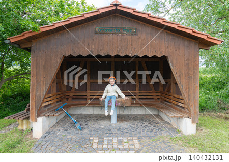 A cute little boy with a denim backpack sitting on a wooden bench inside a rustic rural bus stop shelter. Perfect commercial concept for childhood journeys, countryside travel, and summer holidays A cute little boy with a denim backpack sitting on a wooden bench inside a rustic rural bus stop shelter. Perfect commercial concept for childhood journeys, countryside travel, and summer holidays 140432131