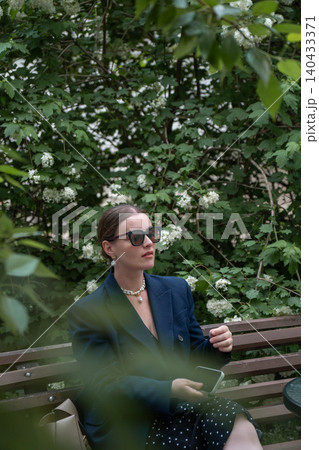 Young woman in sunglasses sits on wooden bench, lush green foliage, white blossoms while holding smartphone in garden setting. Digital lifestyle, mobile communication, remote work, technology branding Young woman in sunglasses sits on wooden bench, lush green foliage, white blossoms while holding smartphone in garden setting. Digital lifestyle, mobile communication, remote work, technology branding 140433371