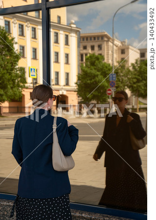 Woman observes her reflection in glass facade in Moscow, historic urban architecture mirrored in modern surface, identity, self-perception, contemporary city life. Fashion content, confidence concept 140433492
