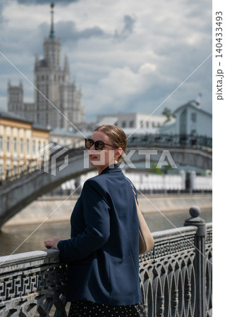 Young woman in dark blazer and sunglasses stands on riverside promenade with Sadovnichesky Bridge and Kotelnicheskaya Embankment Building. Urban travel, city tourism, destination 140433493