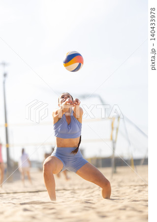 Woman plays beach volleyball, receiving a ball during a match at a sandy court under a bright sun near a sports facility Woman plays beach volleyball, receiving a ball during a match at a sandy court under a bright sun near a sports facility 140433978