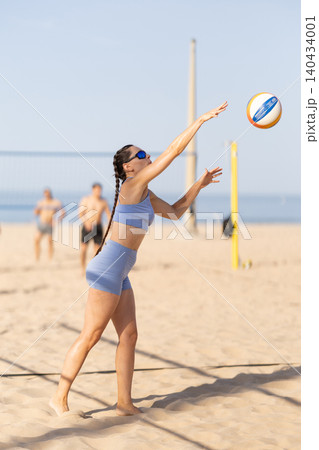 Woman practices volleyball on the beach during a sunny day while others play in the background 140434001