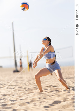 Female player hits volleyball on beach sand during a sunny day with nets and other players in the background 140434020