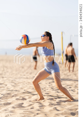 Young woman plays volleyball on sandy beach during sunny day with other players in background Young woman plays volleyball on sandy beach during sunny day with other players in background 140434021
