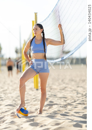 Young woman poses by volleyball net on beach during sunny day while others play in background Young woman poses by volleyball net on beach during sunny day while others play in background 140434025