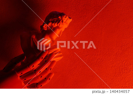 Classical angel statue with praying hands illuminated by dramatic red lighting on textured wall Classical angel statue with praying hands illuminated by dramatic red lighting on textured wall 140434057