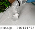Closeup view of bottle pours epoxy resin into clear plastic cup. Hand wearing blue glove holding bottle for safety. Green moss in the background, crafting and handmade process, hobby and leisure 140434758