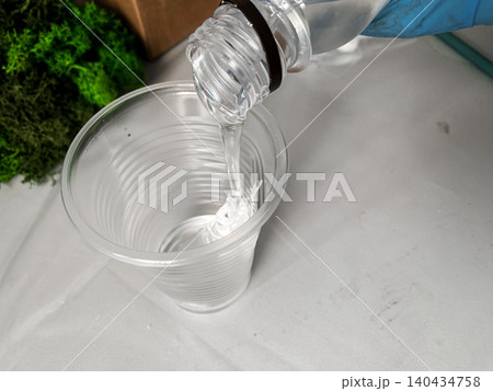 Closeup view of bottle pours epoxy resin into clear plastic cup. Hand wearing blue glove holding bottle for safety. Green moss in the background, crafting and handmade process, hobby and leisure 140434758