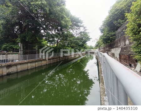 Canal with green water, tree lined banks and concrete wall under cloudy sky Canal with green water, tree lined banks and concrete wall under cloudy sky 140435310