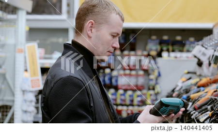 Man in a black jacket carefully selects a cordless drill in a hardware store, carefully comparing features and prices. A man in a hardware store choosing a tool. Man in a black jacket carefully selects a cordless drill in a hardware store, carefully comparing features and prices. A man in a hardware store choosing a tool. 140441405