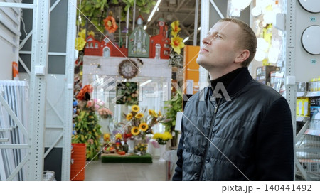 A man in a hardware store, choosing materials for repairs. Man carefully selects lighting for his apartment while standing in a hardware store near the counter and examining products on the shelves 140441492