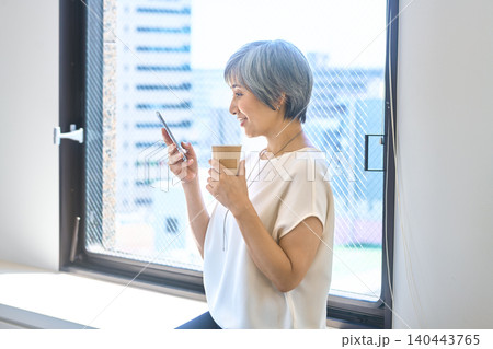 窓辺でスマホを見ながらコーヒーを持つシニア女性 140443765