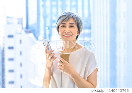 スマホとコーヒーを持って窓辺に立つ笑顔のシニア女性 スマホとコーヒーを持って窓辺に立つ笑顔のシニア女性 140443766