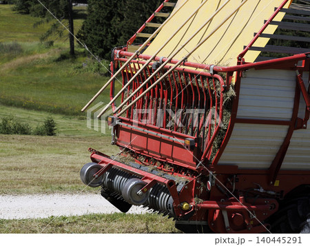Red Forage Wagon Collecting Hay in a Sunny Farm Field During Harvest Season in dolomites 140445291