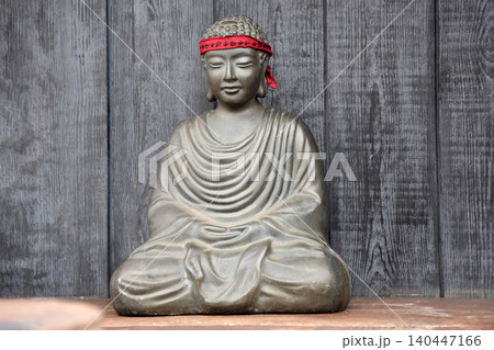 Serene Buddha statue with red headband meditating against wooden backdrop, symbolizing peace and Vesak Serene Buddha statue with red headband meditating against wooden backdrop, symbolizing peace and Vesak 140447166